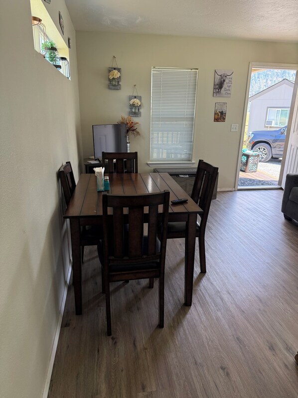 A dining area with a wooden table and chairs, a television, and a window with blinds. - Cozy home on Cattle Ranch, close drive to Crater Lake National Park entrance. (Chiloquin)
