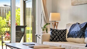 A modern living room with a sofa, a coffee table, and a lamp. There is a window with a view of greenery outside. - Luxury / Beachside / Pickle Ball / Surf / Pool (New Smyrna Beach)