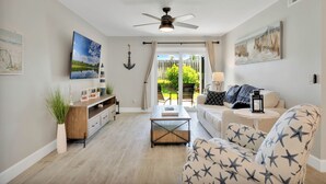 A modern living room with a ceiling fan, a wooden coffee table, a sofa with star patterns, and a view through a glass door to an outdoor area. - Luxury / Beachside / Pickle Ball / Surf / Pool (New Smyrna Beach)
