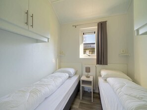 A small, clean room with two single beds, a window with curtains, and a small table with a lamp. - Comfy Chalet With Dishwasher, the Efteling at 2 km (Kaatsheuvel)
