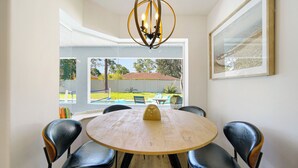 Un comedor con una mesa redonda y cuatro sillas, una araña, un ventanal con vista a un jardín, y un cuadro en la pared.