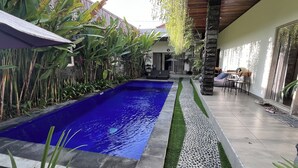 Una piscina con revestimiento azul, rodeada de abundante vegetación y un área de patio con baldosas.