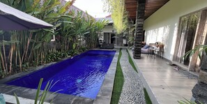 Una piscina con revestimiento azul, rodeada de abundante vegetación y un área de patio con baldosas.