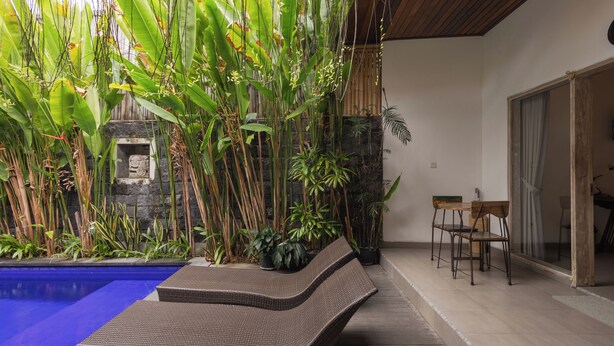 Un área junto a la piscina con sillones de mimbre, un muro de piedra y plantas tropicales.
