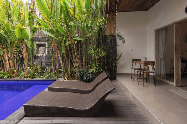 Un área junto a la piscina con sillones de mimbre, un muro de piedra y plantas tropicales.