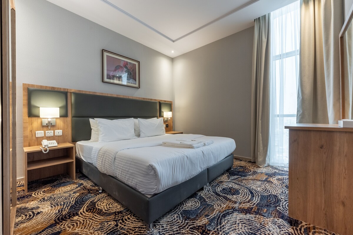 A hotel room with a large bed, a wooden nightstand with a phone, a wall-mounted lamp, and a painting above the bed.
