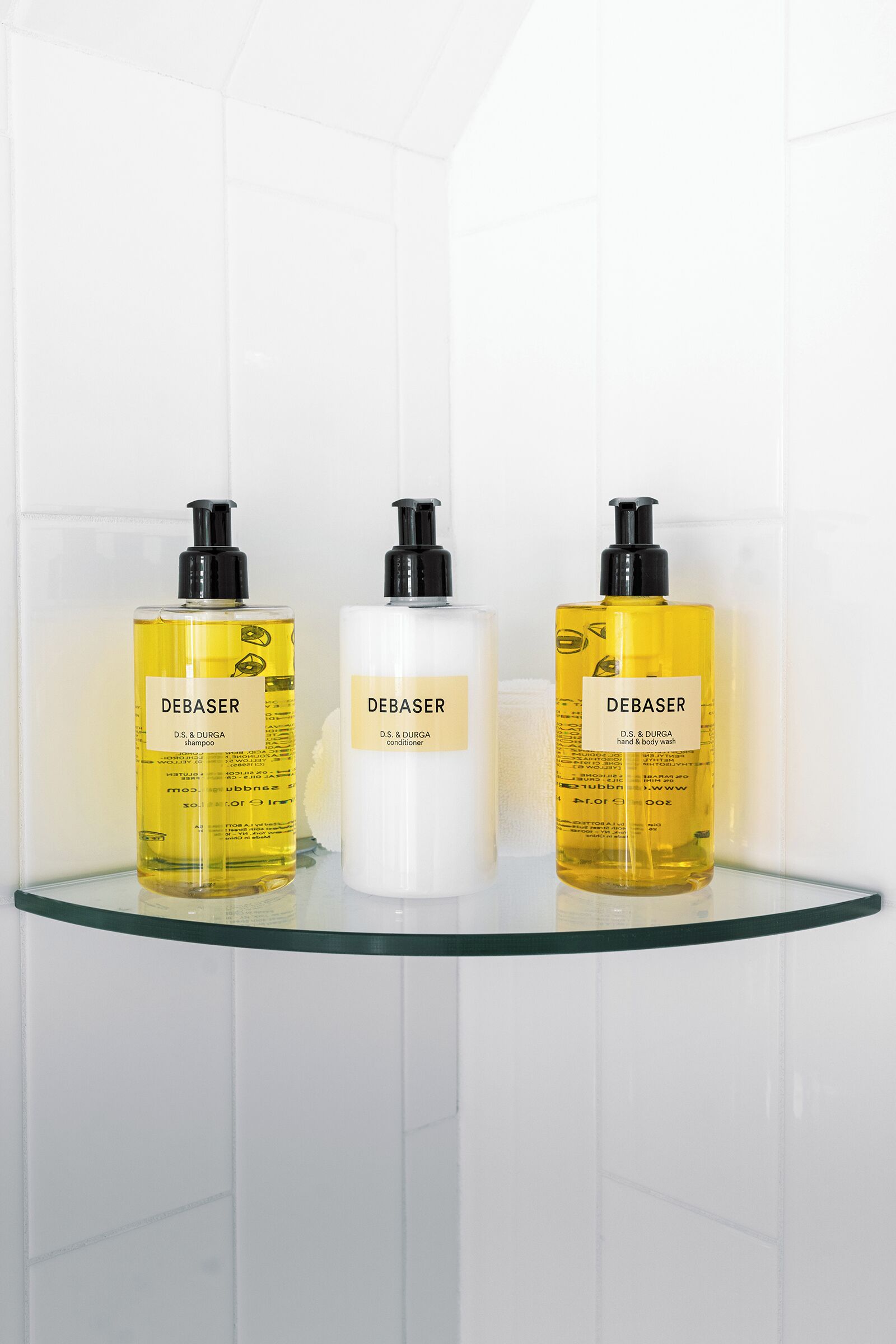 Tres productos Debaser sobre un estante de vidrio frente a una pared de azulejos blancos.