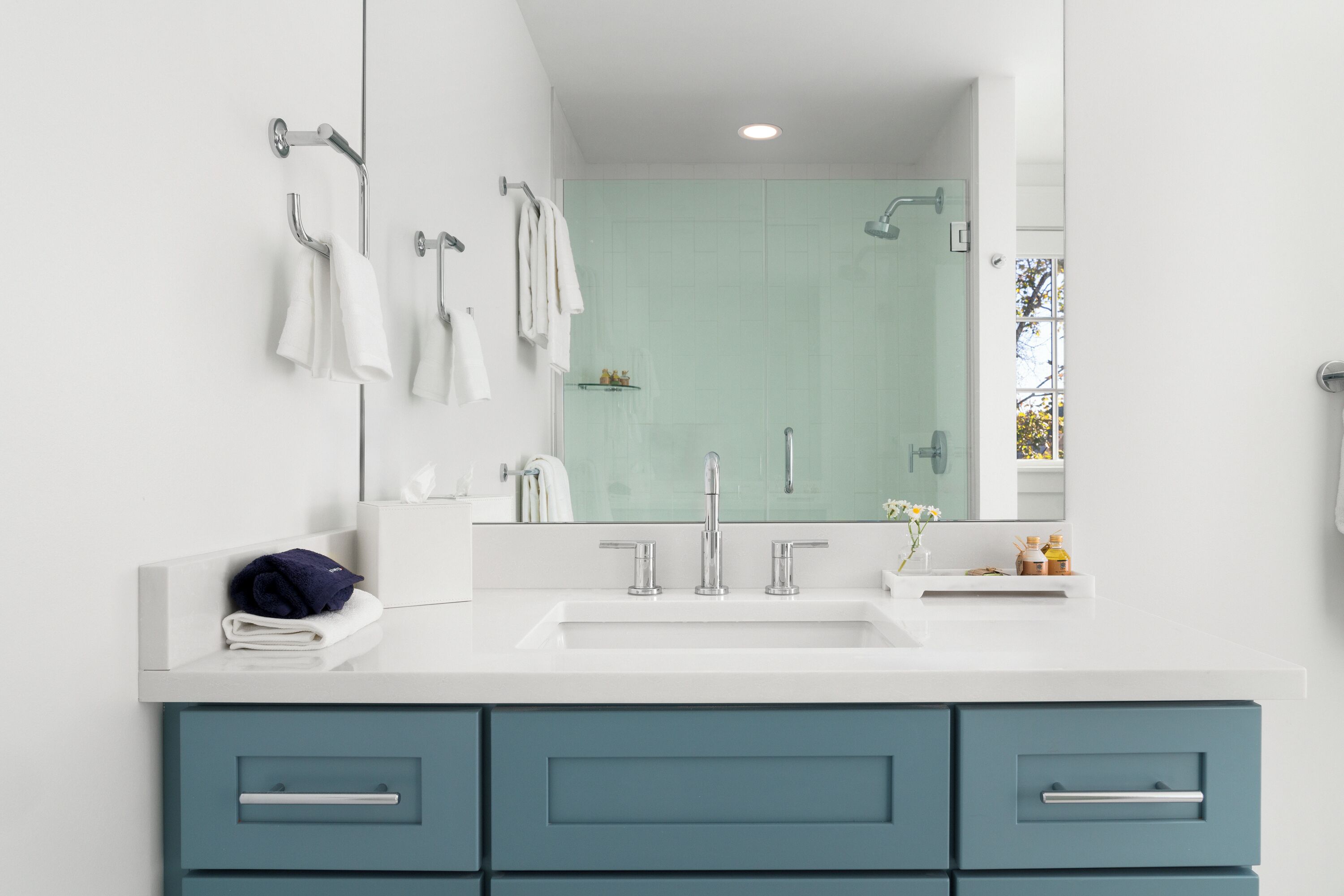Un baño moderno con lavamanos blanco, gabinetes verde azulado y una cabina de ducha de vidrio.