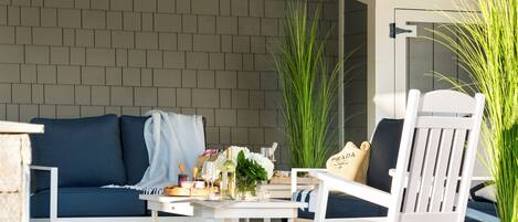 Een terras met een witte tafel, een blauwe bank en witte stoelen. Er staan potplanten en een klein tafeltje met spullen op.