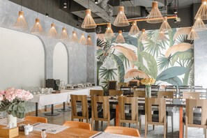 Un restaurante con varias mesas y sillas, lámparas colgantes de bambú y un amplio mural tropical en la pared.