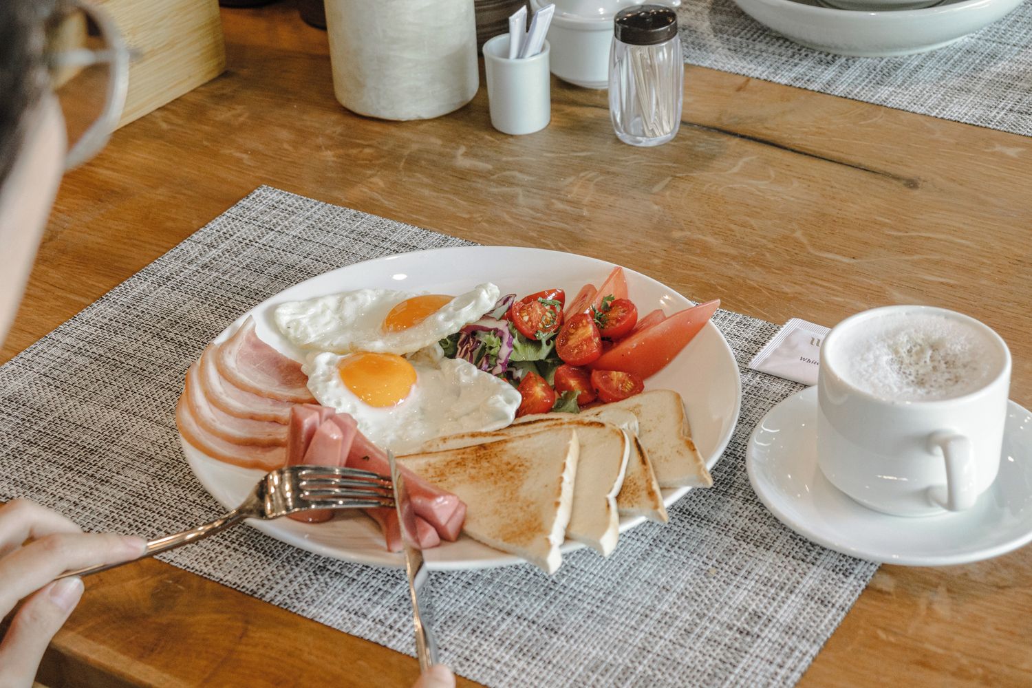 Un plato de desayuno con huevos, tomates y jamón, una taza de café y un tenedor sobre una mesa de madera.
