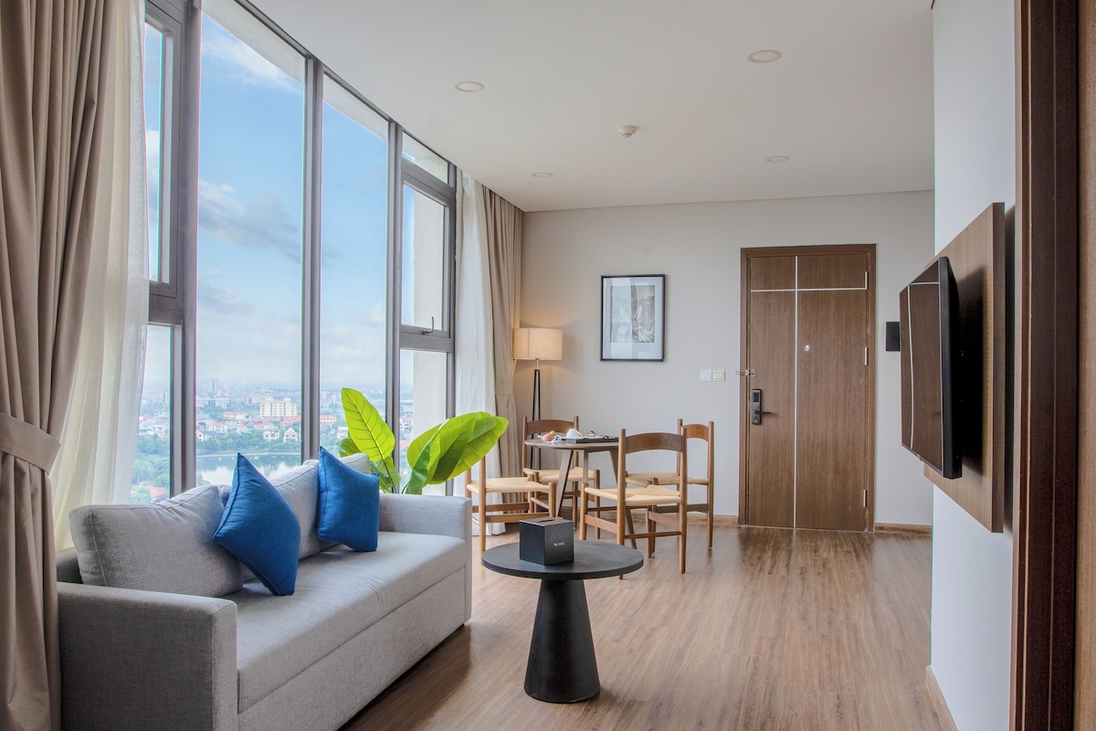 Habitación de hotel moderna con sofá, una mesa de comedor, sillas, televisor y ventanas amplias con vista a la ciudad.