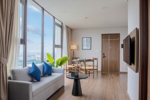 Habitación de hotel moderna con sofá, una mesa de comedor, sillas, televisor y ventanas amplias con vista a la ciudad.