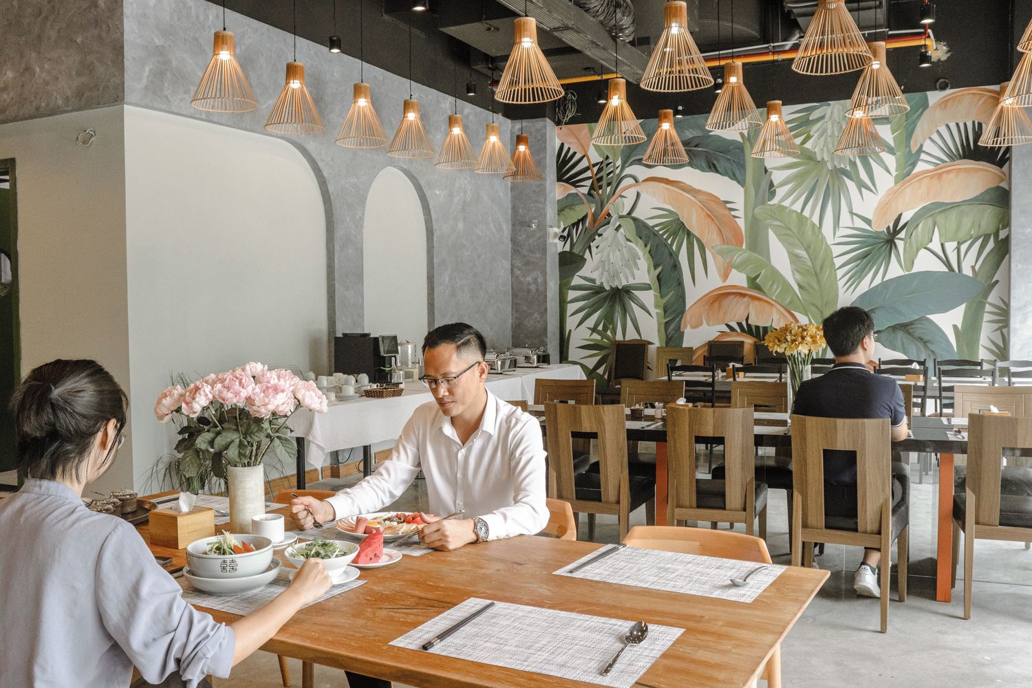 A restaurant with wooden tables and chairs, hanging pendant lights, and a mural of tropical leaves on the wall.