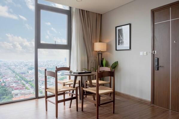 A hotel room with a large window, a round table with chairs, a lamp, and a door. - Mandala Hotel & Suites Hai Duong (Tan Hung Ward, Hai Phong City)