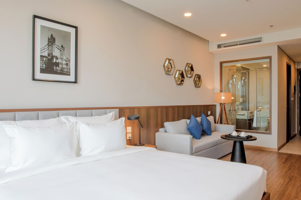 Habitación de hotel con una cama grande, un sofá, una mesita y un baño visible a través de una puerta de vidrio.