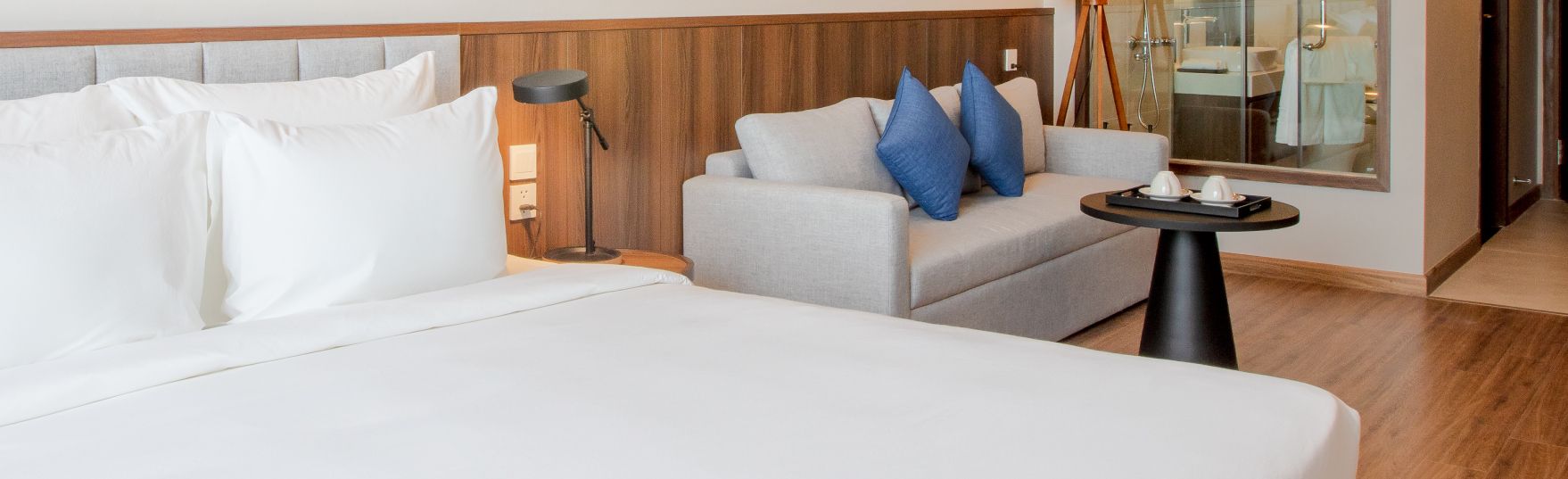 A hotel room with a large bed, a sofa with blue pillows, a small table with a lamp, and a mirror reflecting a bathroom.