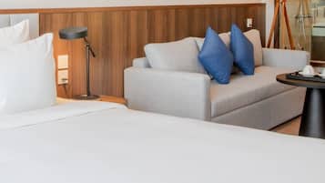A hotel room with a large bed, a sofa with blue pillows, a small table with a lamp, and a mirror reflecting a bathroom.