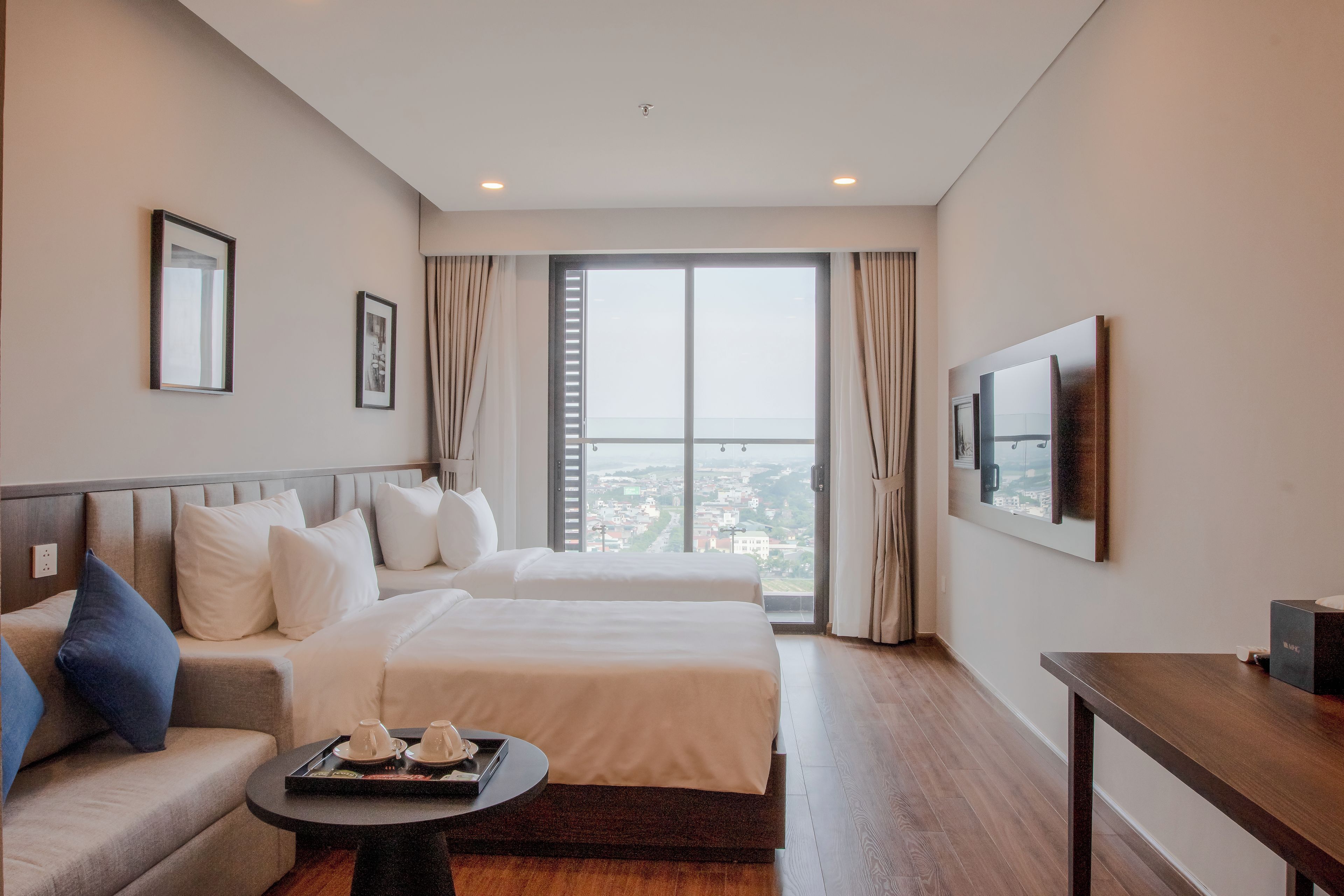 A hotel room with two beds, a sofa, a small table with a tray, a TV, and a large window with a city view.
