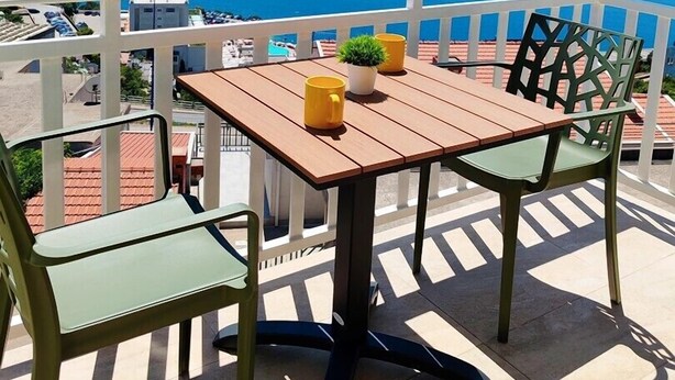 Un balcone con tavolo e sedie in legno, vista su un corpo d'acqua e colline in lontananza.