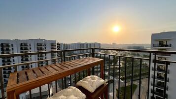 A balcony with a wooden table and two cushioned seats, overlooking a cityscape with multiple buildings and a clear sky.