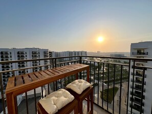 A balcony with a wooden table and two cushioned seats, overlooking a cityscape with multiple buildings and a clear sky.