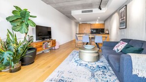 Un salon moderne avec un canapé, une table basse et de grandes plantes. On aperçoit un coin repas avec des chaises et, au fond, une cuisine.