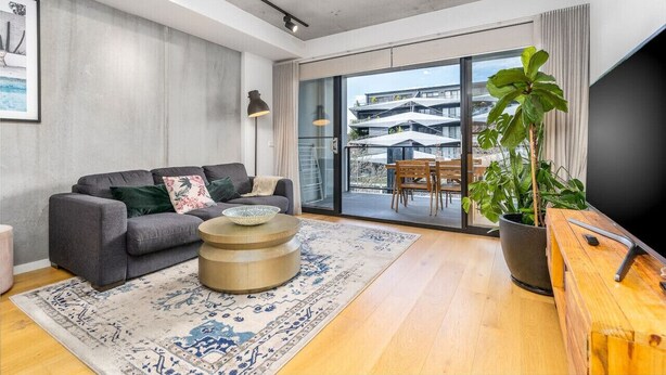 Un salon moderne avec un canapé gris, une table basse ronde, une grande plante en pot et une vue sur un immeuble à travers de grandes fenêtres.
