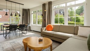 Een moderne woonkamer met een bank, twee houten tafels, een eethoek met stoelen, en grote ramen met uitzicht op groen en huizen.
