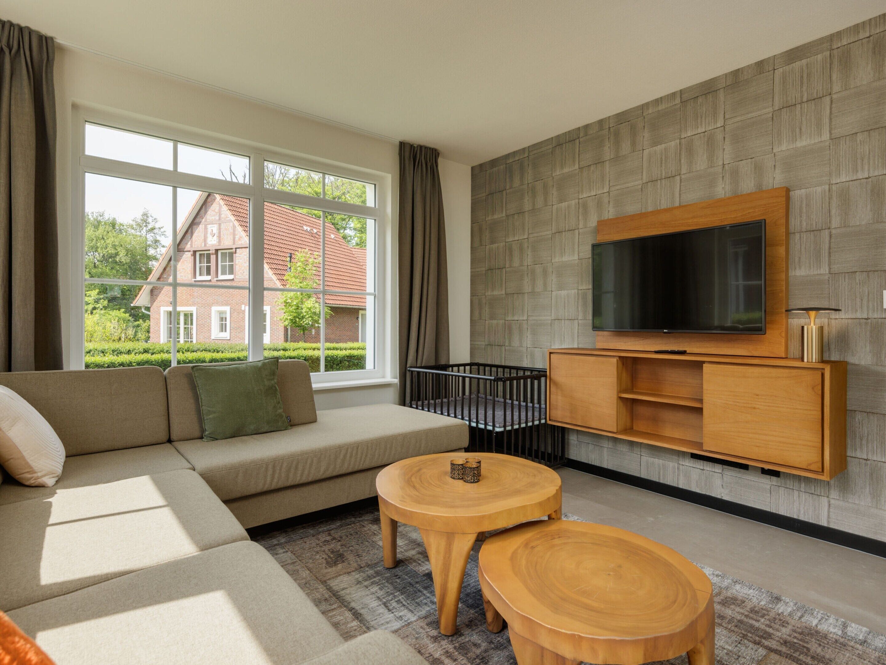 A modern living room with a sofa, two wooden side tables, a television on a wooden cabinet, and a view of a house through large windows.