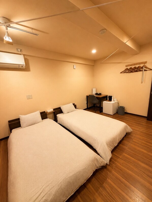 A hotel room with two beds, a desk, a chair, a small table, an air conditioning unit, and a hanging clothes rack. - Jinya Sunset Inn by Murasaki (Takayama)