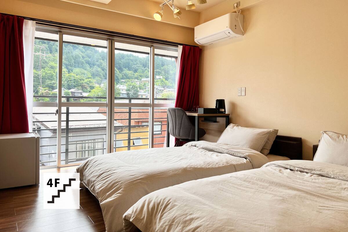 Une chambre d’hôtel avec deux lits, un bureau avec une chaise, un climatiseur et une grande fenêtre avec des rideaux rouges.