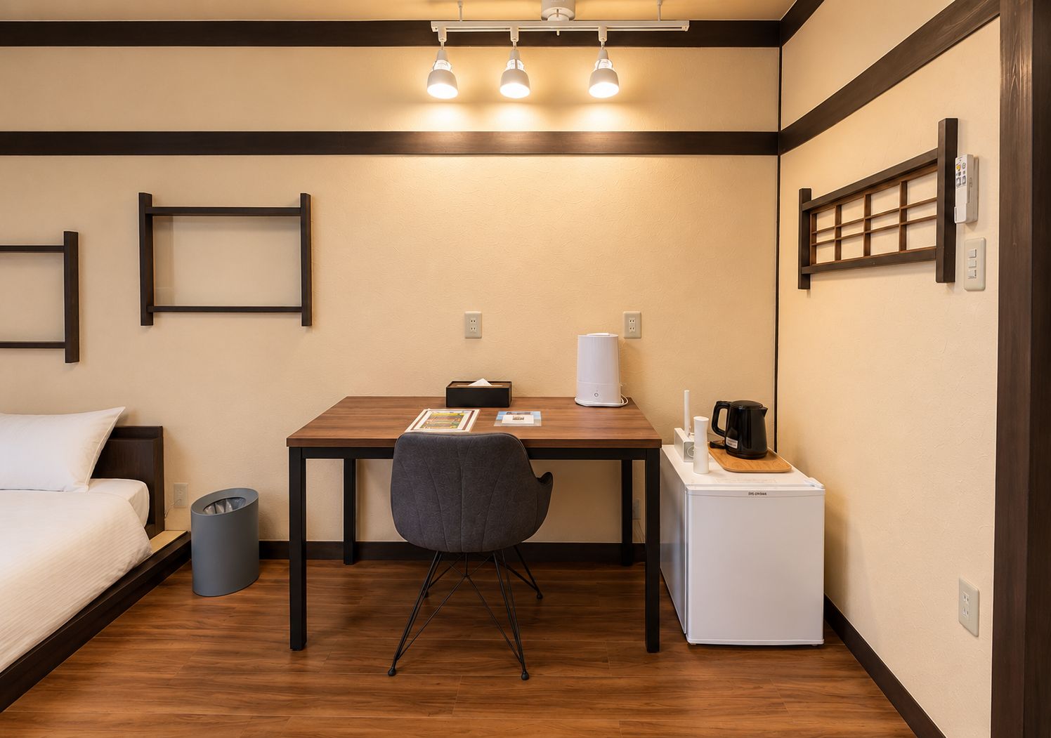 A compact hotel room with a single bed, a small desk with a chair, a coffee maker, a mini fridge, and a trash bin.