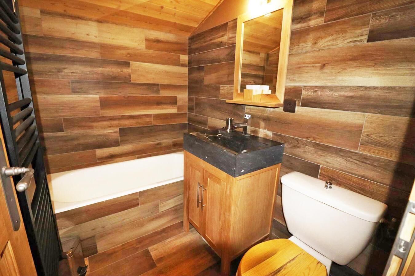 A bathroom with a wooden floor, a black countertop sink, a wooden cabinet, a white toilet, and a wooden-framed mirror.