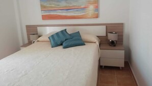 Ein Schlafzimmer mit einem Bett, zwei Kissen, einem Nachttisch mit Lampe und einem Gemälde von einem Sonnenuntergang über dem Meer an der Wand.