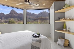 Une chambre dotée d’un grand lit, de deux fenêtres donnant sur la montagne, d’un ventilateur de plafond et d’étagères intégrées ornées d’objets décoratifs.