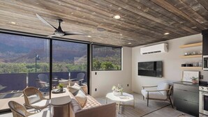 Un intérieur moderne avec un plafond en bois, de grandes fenêtres offrant une vue sur les montagnes, un coin repas avec des chaises en osier, un canapé et une cuisine équipée d’un téléviseur et d’appareils électroménagers.