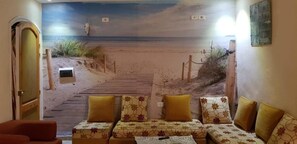 A room with a beach mural, a wooden door, and a sofa with floral cushions. - Douja Apartment One-Bedroom Apartment (NABEUL CAPBON)