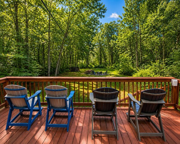 A deck with four Adirondack chairs, a fire pit, and a view of a forested area.
