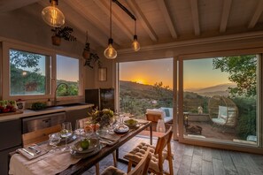 Comedor con una mesa preparada para cuatro personas, ventanas amplias que ofrecen vistas al atardecer, y lámparas colgantes.