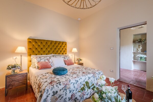 Een slaapkamer met een gestoffeerd hoofdeinde, een bloemenbedovertrek, twee bedlampjes en een nachtkastje met een bloevas.