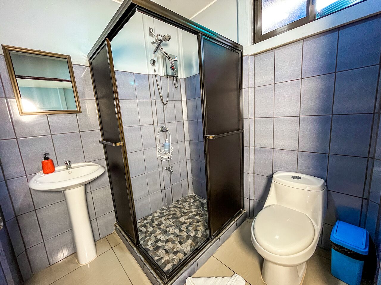 A bathroom with a glass-enclosed shower, a pedestal sink, a toilet, and a blue trash bin.