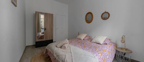 A bedroom with a bed, a wardrobe, a round mirror, and a small table with a lamp.