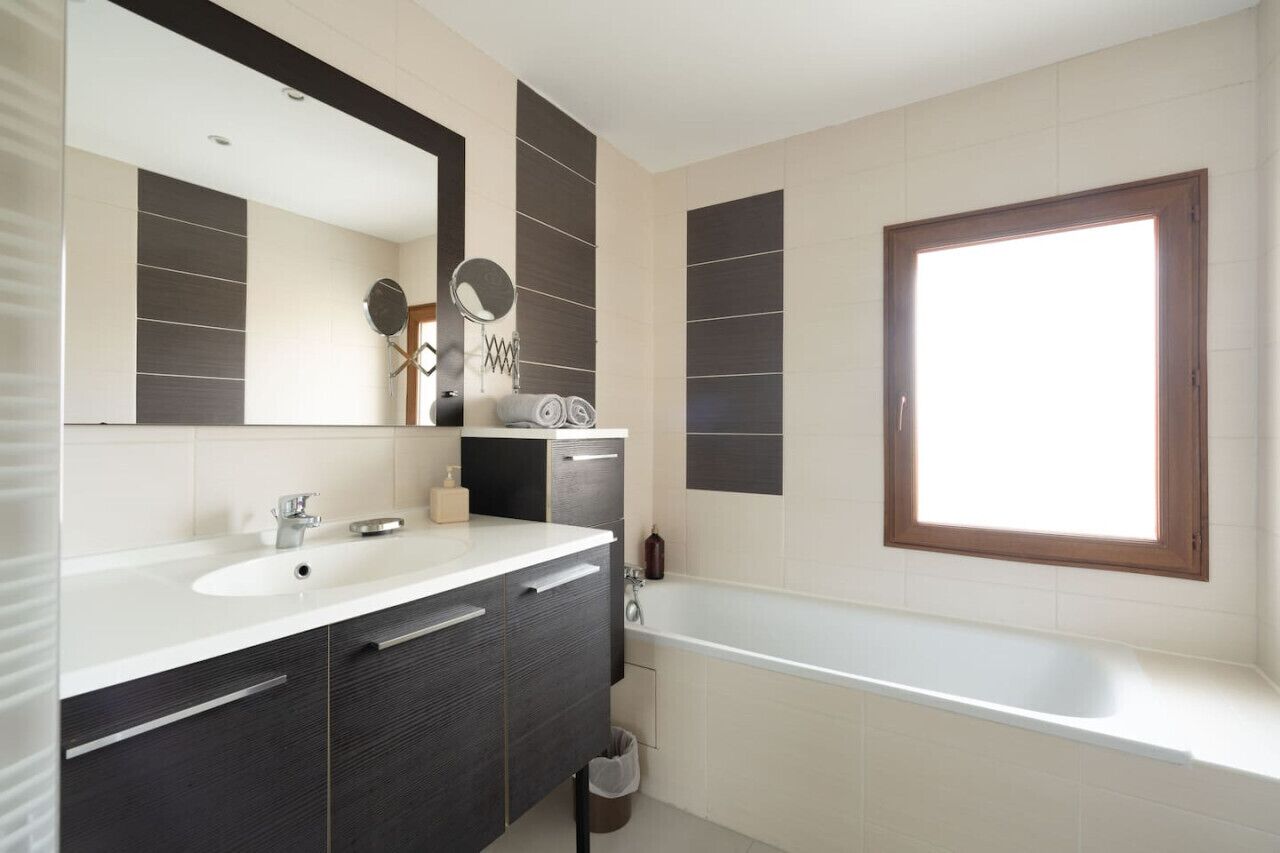 A modern bathroom with a large mirror, a bathtub, and a window.