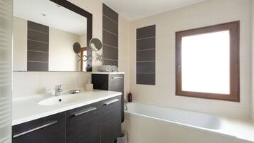 A modern bathroom with a large mirror, a bathtub, and a window.