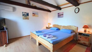 A bedroom with a wooden bed, two pillows, a nightstand with a lamp, a wall clock, and a TV on a stand.