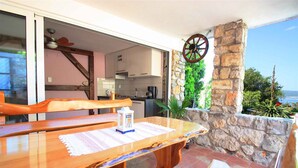 A wooden table with a placemat, a ceiling fan, a stone wall, and a view of the sea.