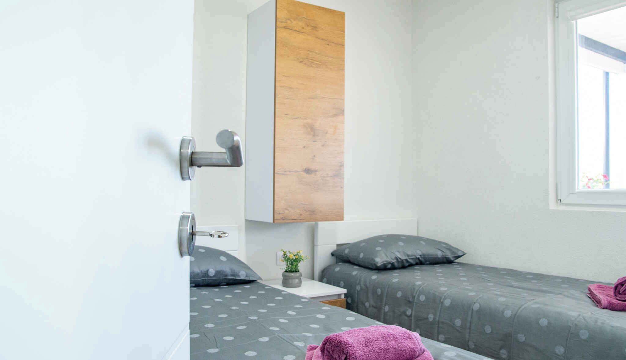 A small, modern bedroom with two single beds, a wooden panel wall, a small bedside table with a vase of flowers, and a window providing natural light.