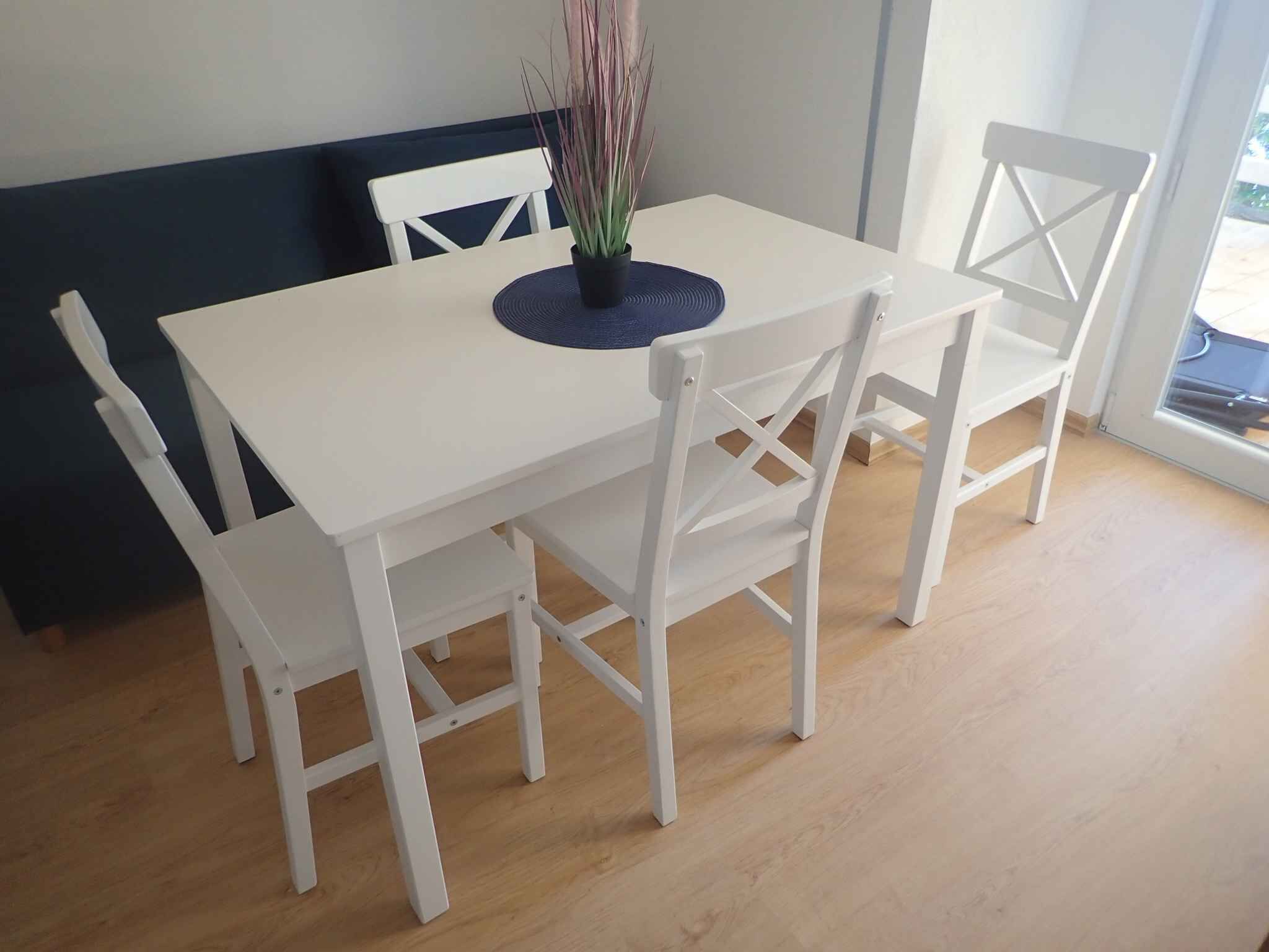 A white dining table with four chairs, a potted plant, and a dark blue placemat.