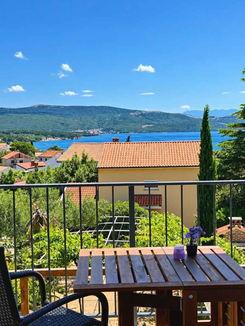 Un balcone con tavolo e sedie in legno, affacciato su un paesaggio costiero con case e un corpo d'acqua.